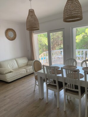 A modern living room with a white sofa, dining table with white chairs, wicker pendant lights, and a view of greenery outside. - Maison à la Mer 8 pers. 60 kms Perpignan (CT)