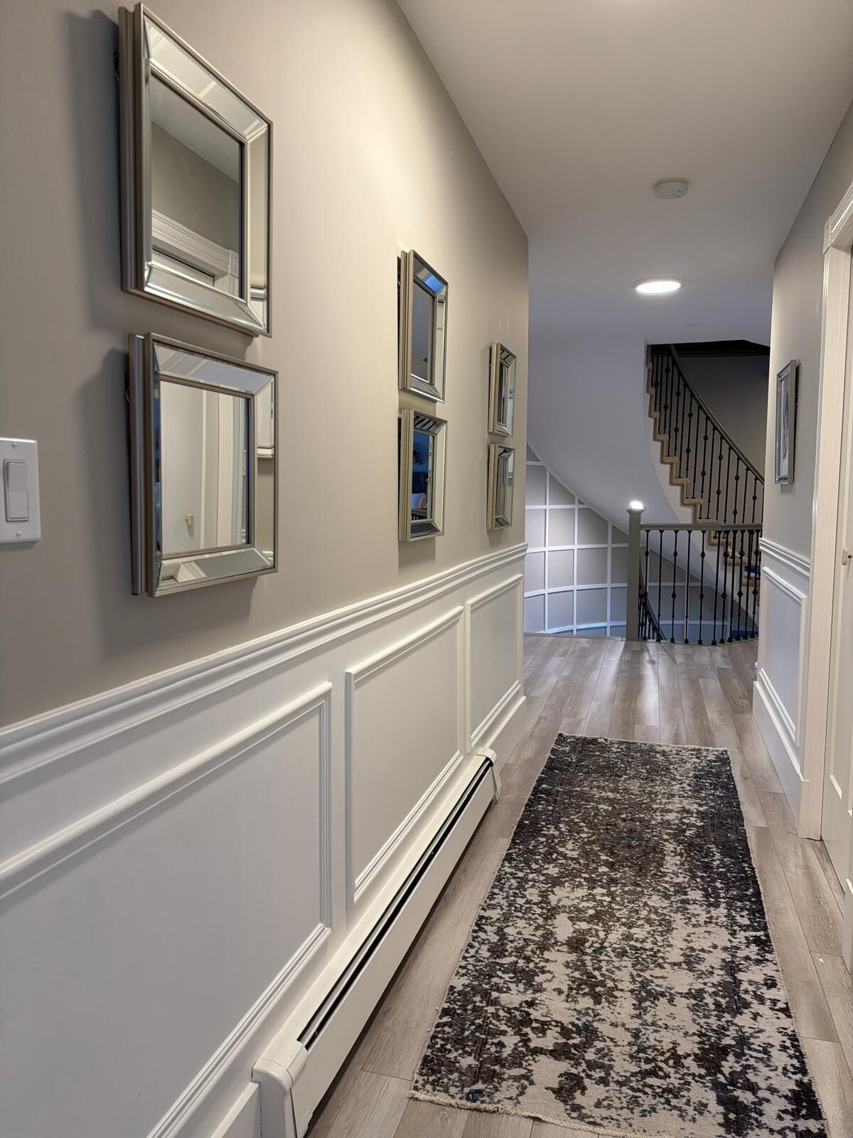A hallway with a white wainscoting, a patterned rug, and a staircase with a railing.