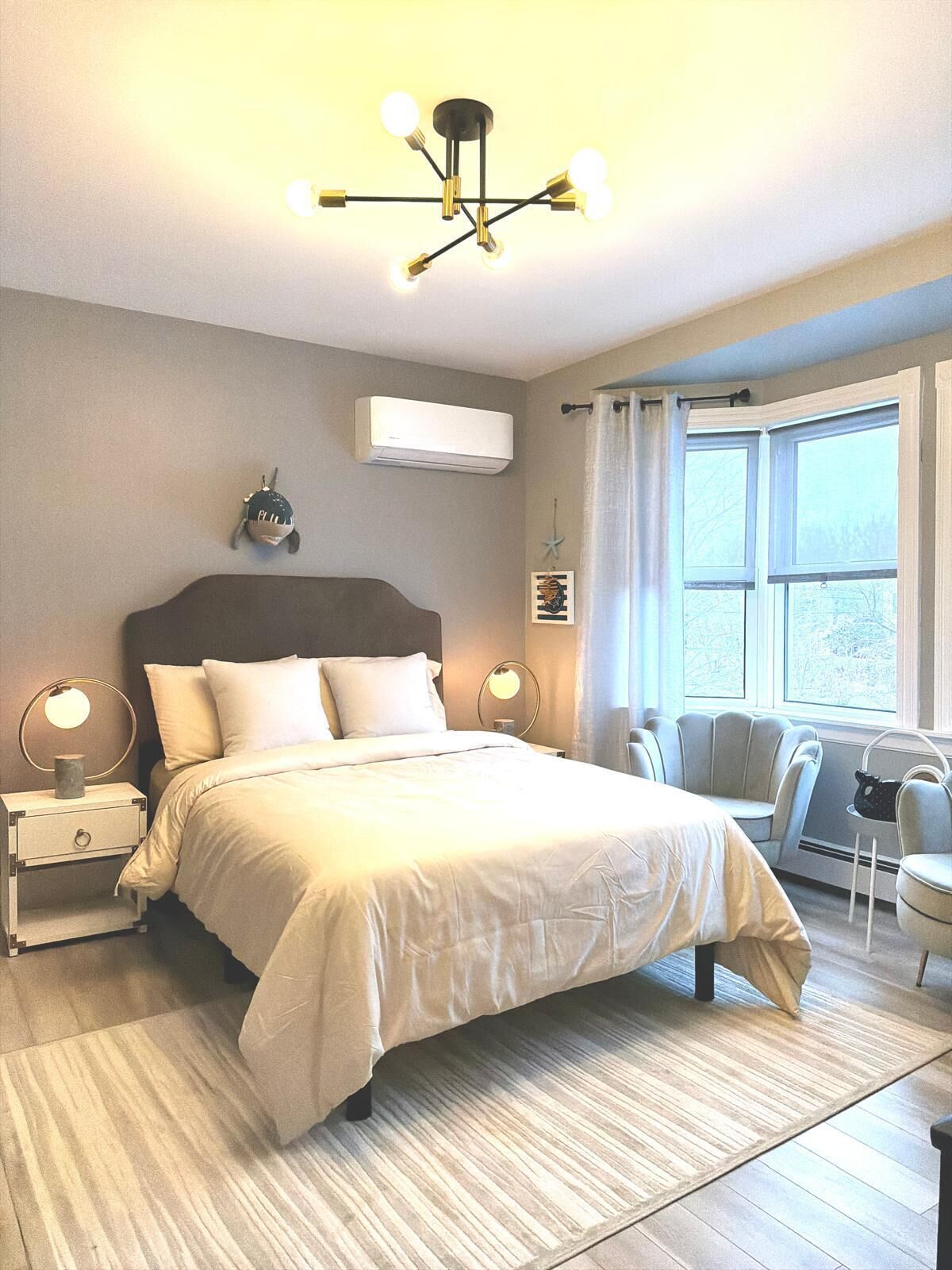Una camera da letto con un letto, comodini, condizionatore d'aria a parete, una finestra con tende e una sedia.