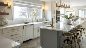Een moderne keuken met witte kasten, een groot eiland met krukken, een inbouwoven en een raam dat voor natuurlijk licht zorgt.