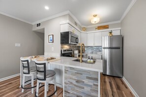 A modern kitchen with white cabinetry, stainless steel appliances, and a central island with stools. - Heron’s Reserve | Oceanfront + Beach Access (North Topsail Beach)