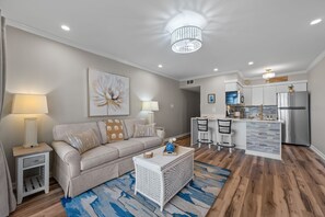 A modern living room with a sofa, coffee table, and a kitchen area with a refrigerator and island. - Heron’s Reserve | Oceanfront + Beach Access (North Topsail Beach)