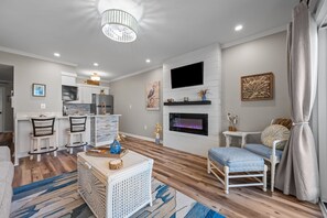 A modern living room with a kitchen, a fireplace, and a TV mounted on the wall. - Heron’s Reserve | Oceanfront + Beach Access (North Topsail Beach)
