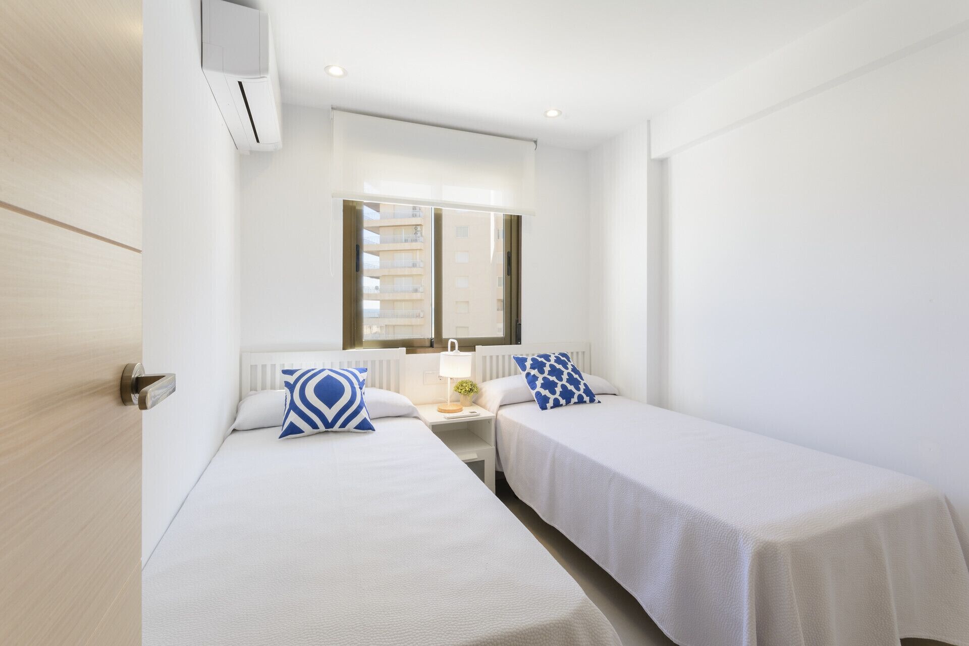 A hotel room with two single beds, a window with a view of buildings, a bedside table with a lamp, and a wall-mounted air conditioner.