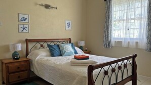 A bedroom with a wooden bed, two bedside tables, a nightstand, a window with sheer curtains, and wall art. - 3-bedroom home away from home near Oistins (Christ Church)