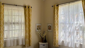 A bedroom with a bed, two windows with curtains, a small table with a vase, and framed pictures on the wall. - 3-bedroom home away from home near Oistins (Christ Church)