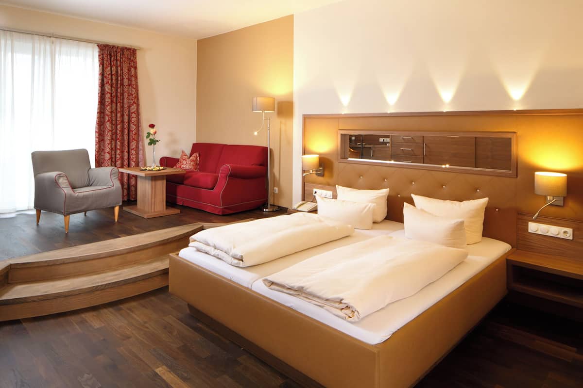 A hotel room with a large bed, a red armchair, a grey armchair, a small table with a vase of flowers, and a lamp.