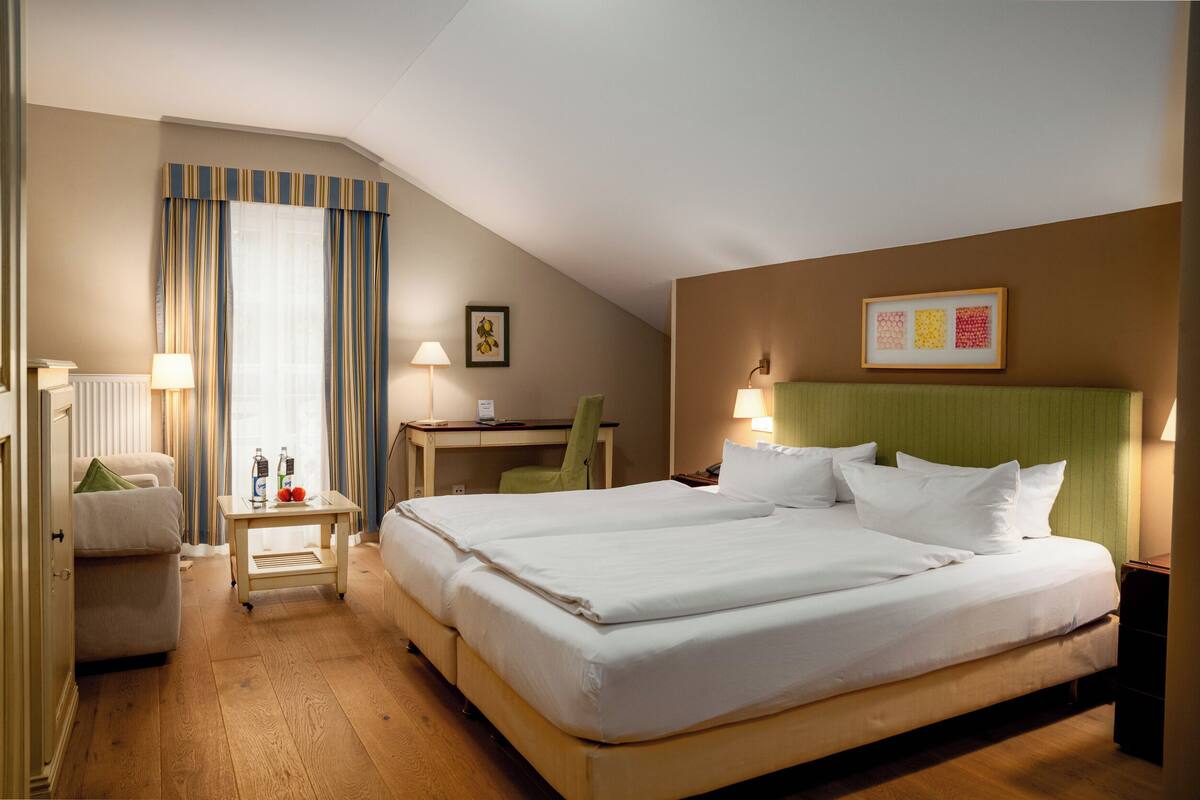 A hotel room with a large bed, a desk, a chair, a small table with bottles, and a radiator.
