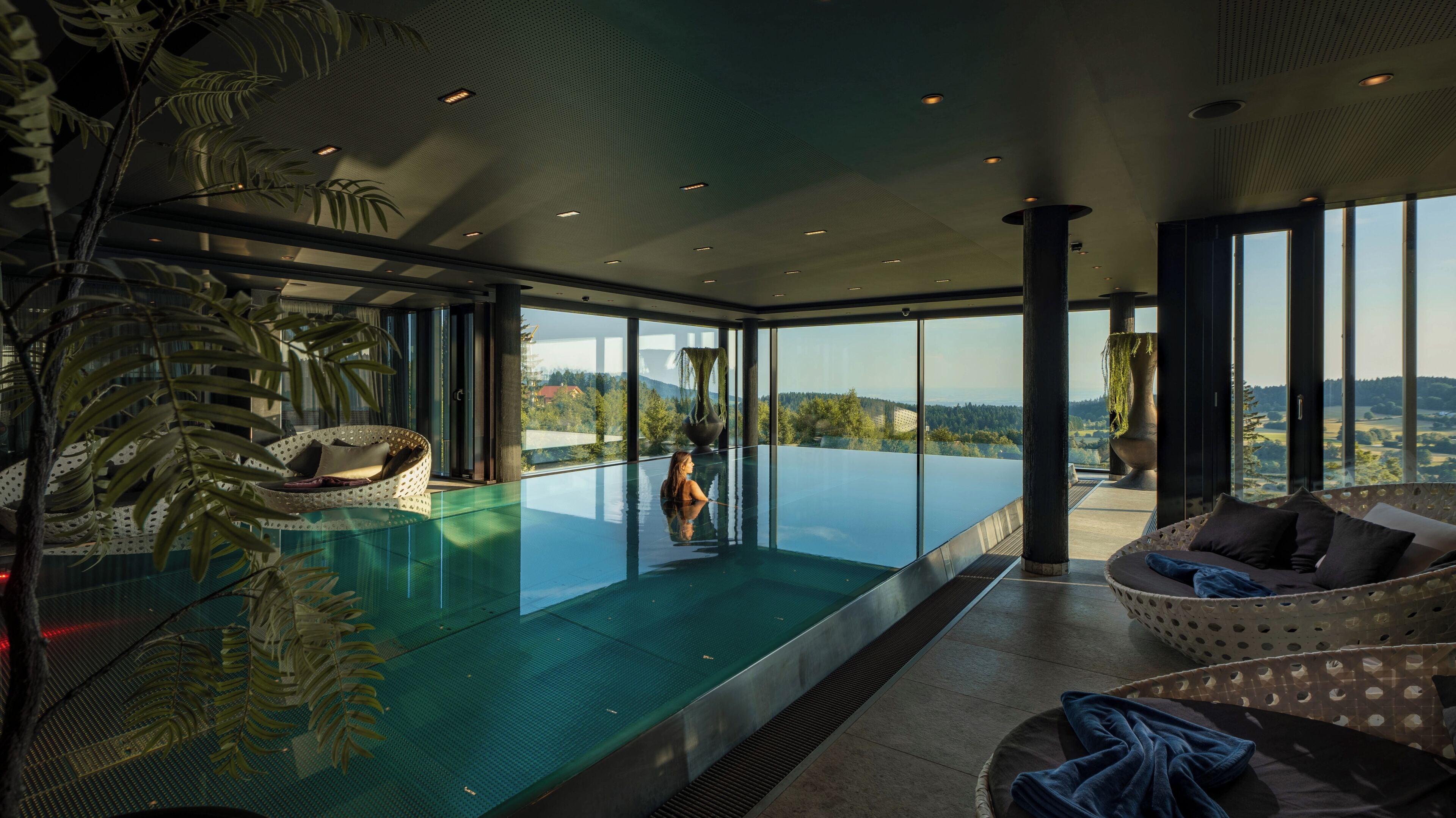 A modern infinity pool with a view of the landscape, a lounge area with a sofa, and a large window.
