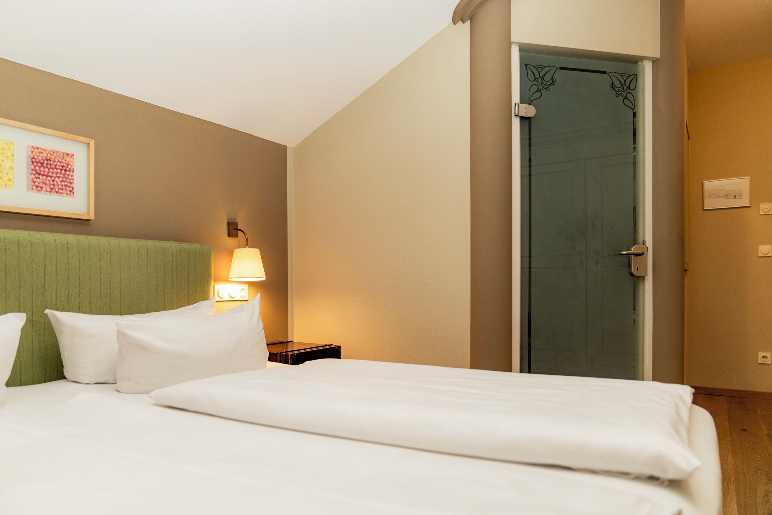 Una habitación de hotel con una cama, almohadas, una mesita de noche, una lámpara, un cuadro en la pared y una puerta.
