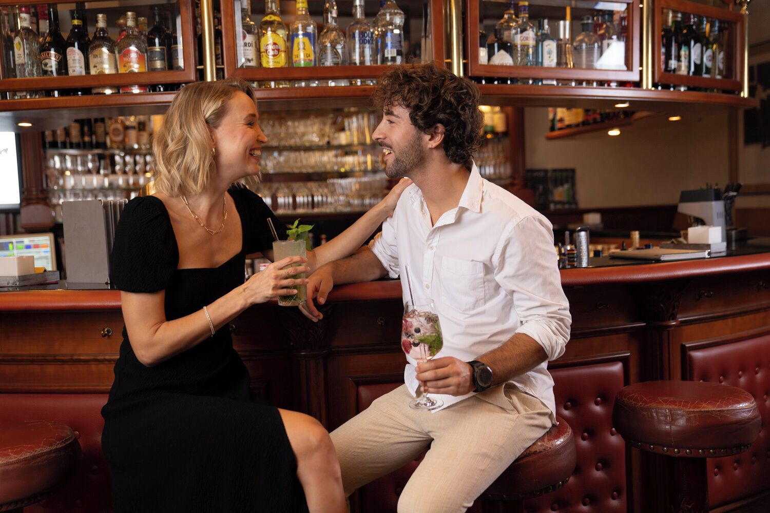 Un bar con una persona sosteniendo una bebida, otra sentada, y una barra repleta de botellas.