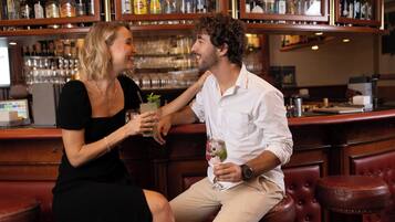 Un bar con una persona sosteniendo una bebida, otra sentada, y una barra repleta de botellas.