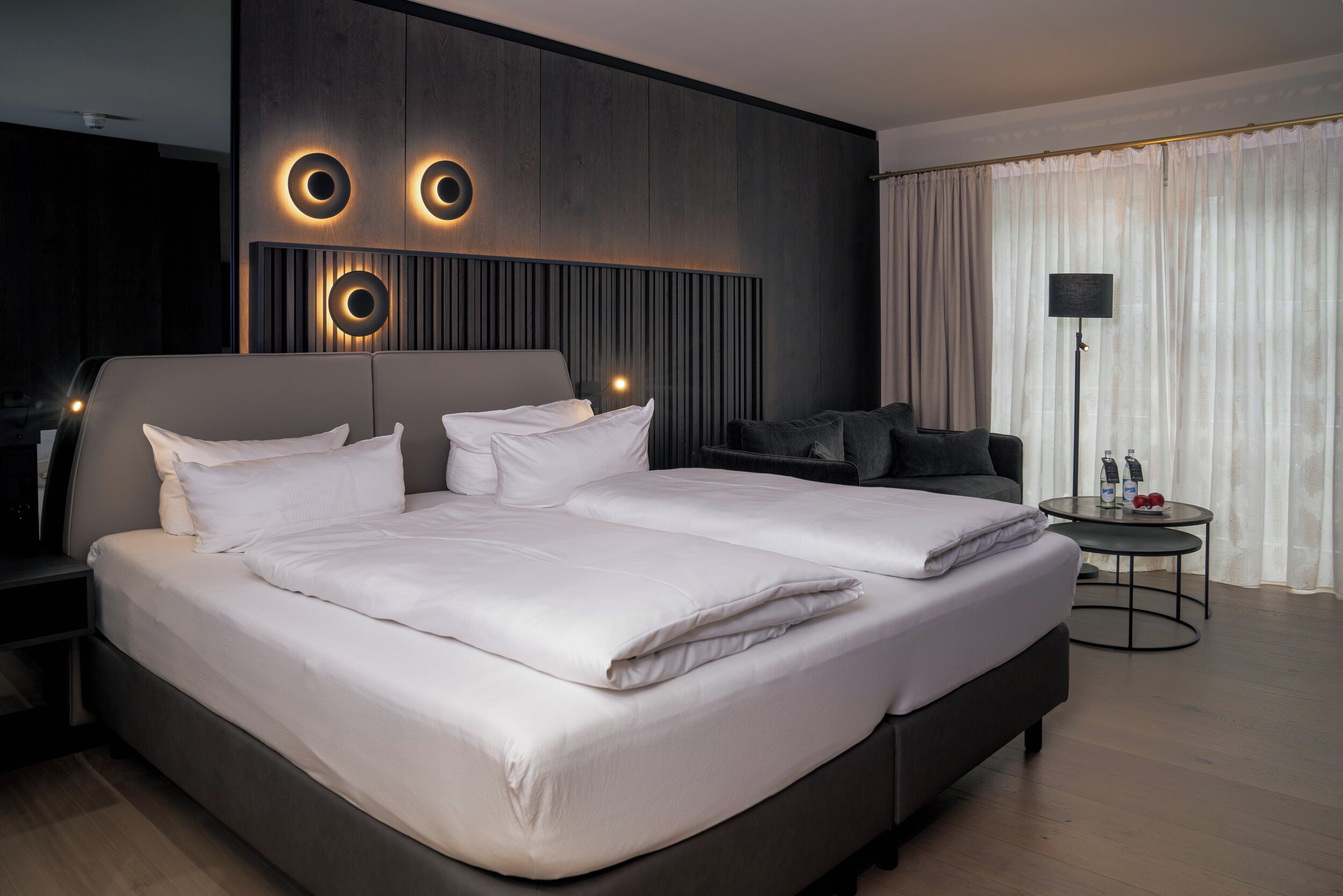 Una habitación de hotel moderna con una cama grande, un sofá, una mesita con una lámpara y un televisor montado en la pared.
