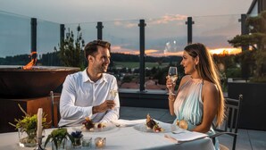 Una pareja cenando al aire libre con vistas a una puesta de sol y una fogata al fondo.
