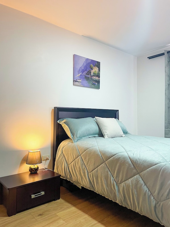 A bedroom with a bed, a nightstand with a lamp, a wall-mounted painting, and a wooden floor.