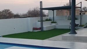 Una piscina con borde de baldosas, pared blanca y estructura de pérgola.