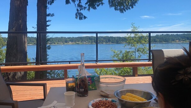 A table with food items and a bottle, overlooking a lake with trees and a clear sky.