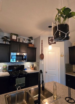 A kitchen with a stainless steel sink, a microwave, and a stove. There is a hanging plant, a door, and a cabinet with decorative items.