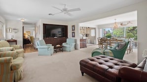 Een ruime woonkamer met een grote flatscreen-tv, meerdere zitmogelijkheden waaronder een bruine leren bank, lichtblauwe fauteuils en groene stoelen.
