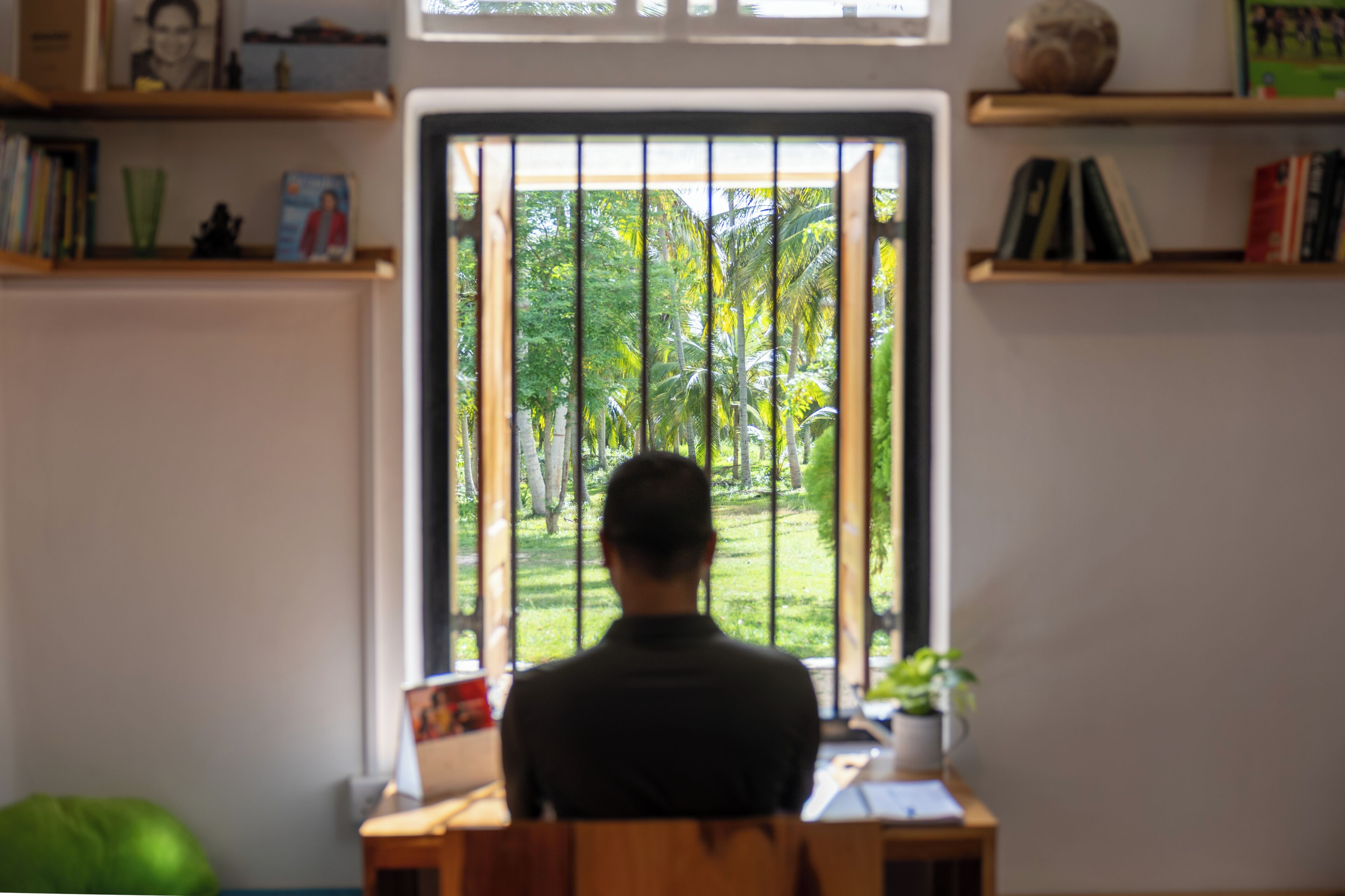 A person seated at a desk facing a window with vertical blinds, overlooking a garden with trees and grass.