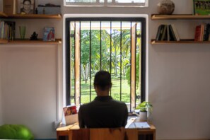 A person seated at a desk facing a window with vertical blinds, overlooking a garden with trees and grass.