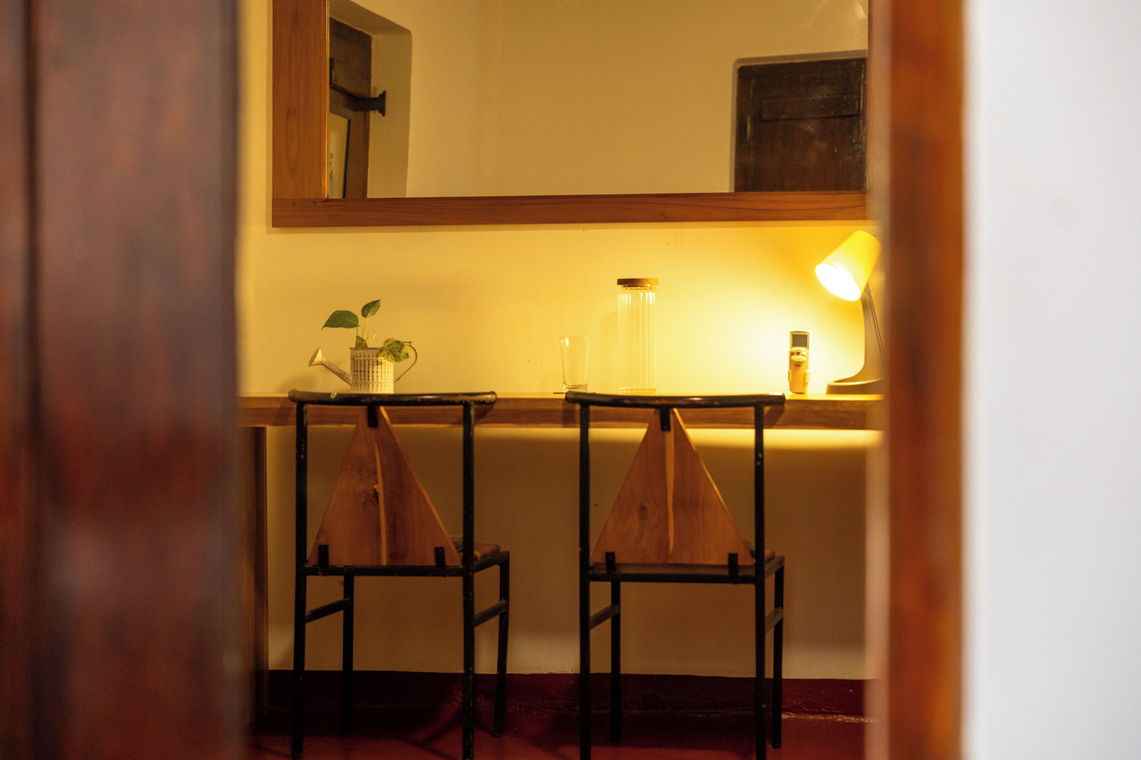 A room with a wooden-framed table and two chairs, a mirror above the table, and a lamp on the table.