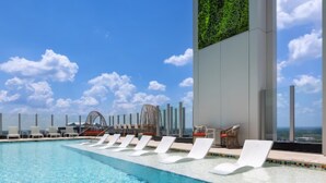 Una piscina con sillones reclinables, barandilla de vidrio y vistas al cielo y nubes.
