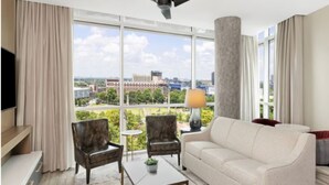 Una habitación de hotel moderna con un ventanal grande, un sofá, dos sillones, una mesita de centro y un televisor.