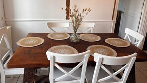 Una mesa de comedor con seis sillas blancas, un florero con plantas secas y cuatro individuales.