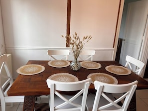 A dining table with six white chairs, a vase with dried plants, and four placemats.