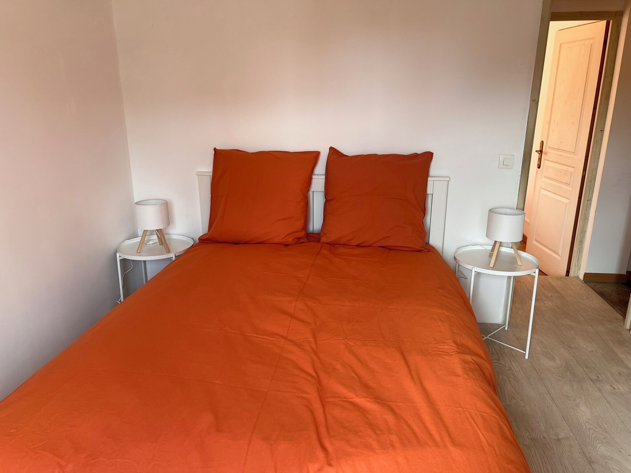 Una cama individual con colcha y almohadas naranja, dos mesitas de noche con lámparas, y una puerta de madera al fondo.
