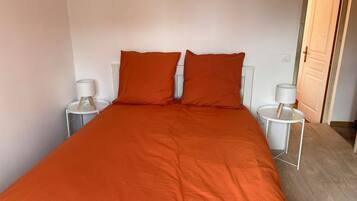 Una cama individual con colcha y almohadas naranja, dos mesitas de noche con lámparas, y una puerta de madera al fondo.