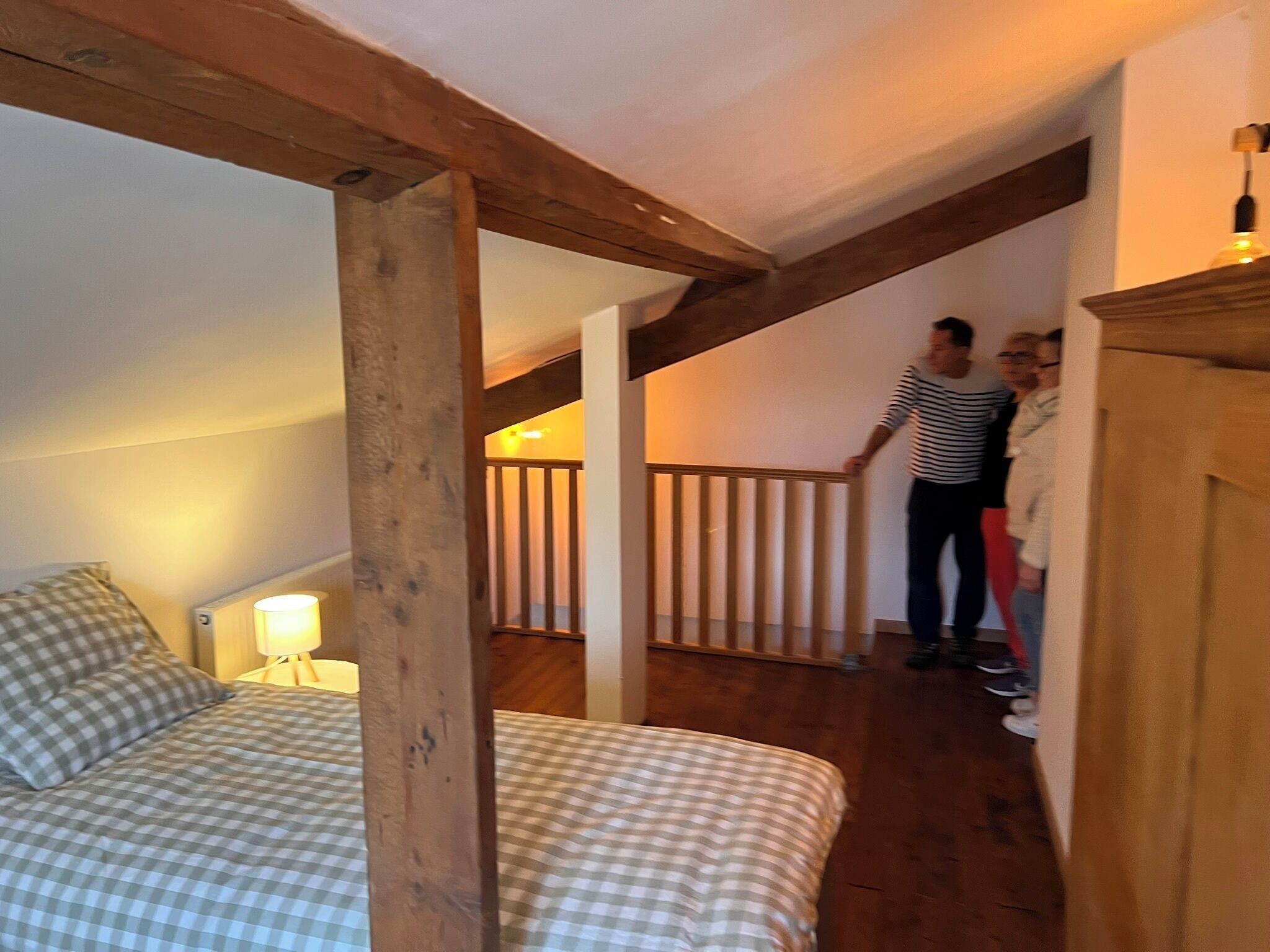 Ein Schlafzimmer mit karierter Bettdecke, Holzbalken, einem Nachttisch mit Lampe und einer Treppe, die zu einer oberen Ebene führt.