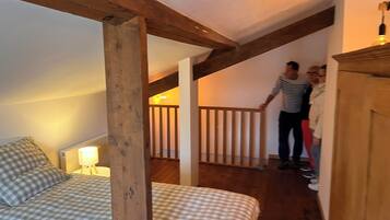 Ein Schlafzimmer mit karierter Bettdecke, Holzbalken, einem Nachttisch mit Lampe und einer Treppe, die zu einer oberen Ebene führt.