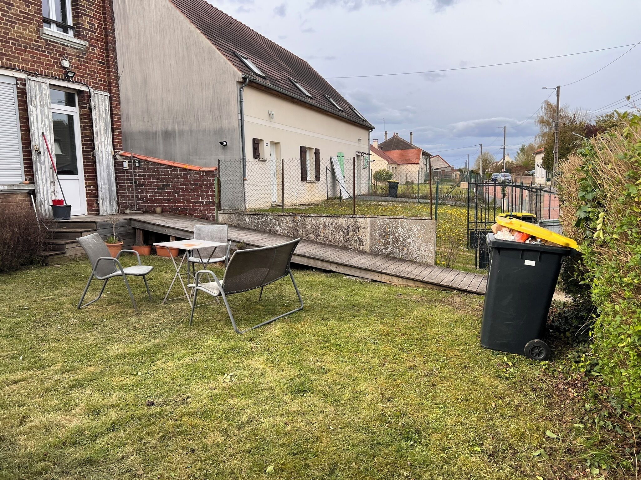 En bostadsträdgård med en uteplats, stolar, ett bord, en soptunna och ett staket.