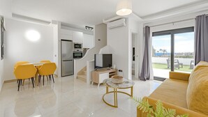 Ένα σύγχρονο σαλόνι με έναν καναπέ, ένα τραπέζι τραπεζ αρίας με καρέκλες, μια τηλεόραση και μια κουζίνα με οικιακές συσκευές.