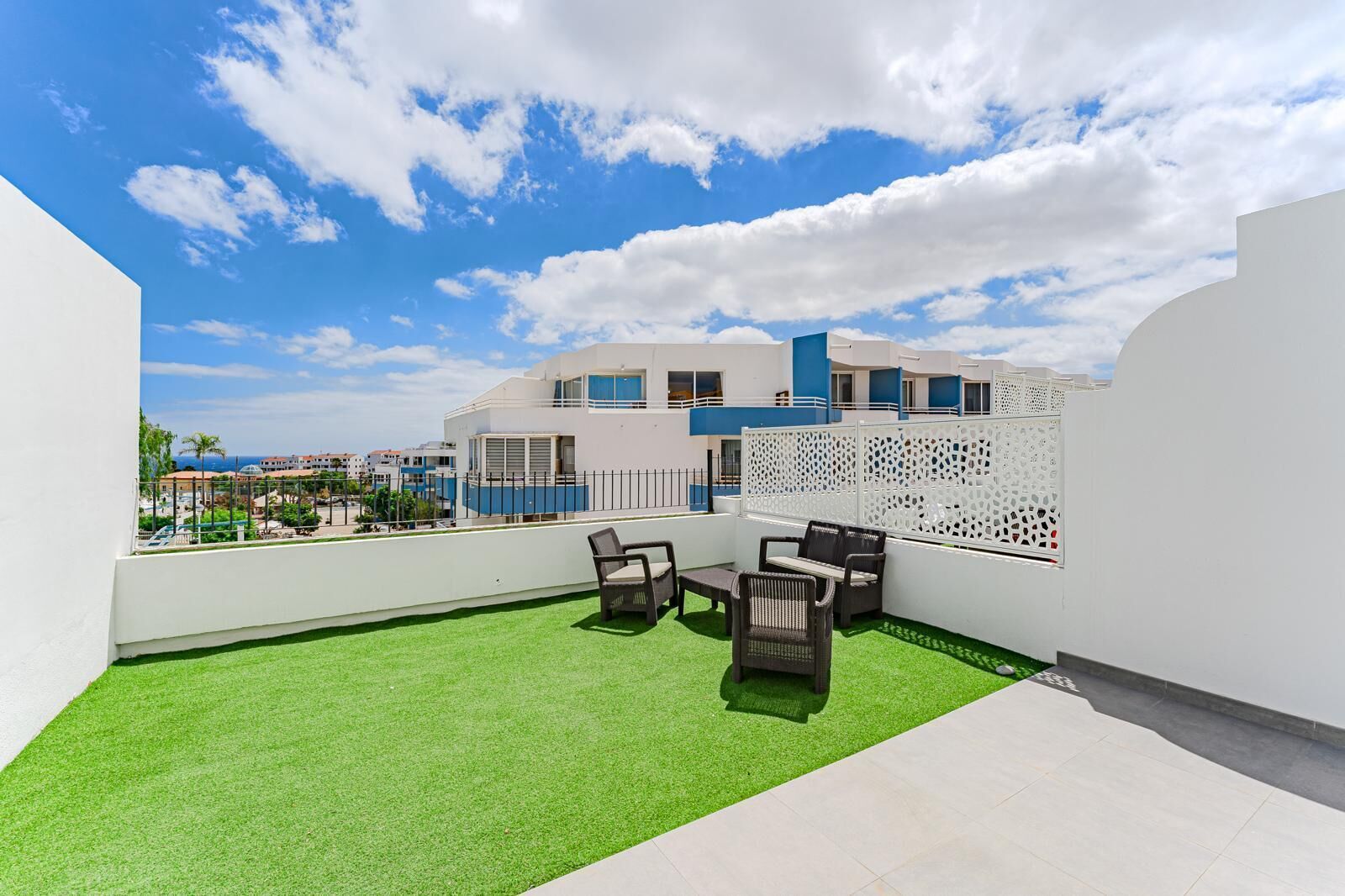 A rooftop terrace with artificial grass, outdoor furniture, and a view of a modern building complex.
