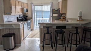 A kitchen with wooden cabinets, a stainless steel trash bin, a refrigerator, a microwave, a coffee maker, and a bar stool area. - Perfect 4‑Bedroom Beach House; Short Walk to the Ocean & Minutes to Great Golf (Myrtle Beach)