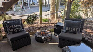 Twee rieten stoelen met kussens waarop 'BEACH HOUSE' en 'RELAX' staat, een kleine vuurplaats van natuursteen en een helder uitzicht op een straat en gebouwen op de achtergrond.