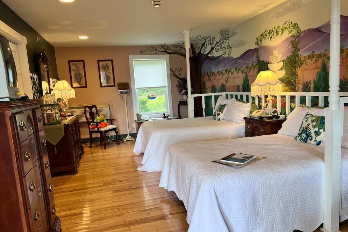 A room with two beds, wooden floors, a mural on the wall, and a window with blinds.