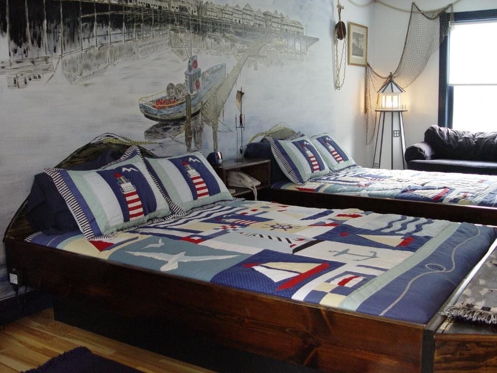 Een kamer met een groot bed, voorzien van een quilt met vliegtuig- en vlagpatronen, een muurschildering van een kustlandschap en een lamp.