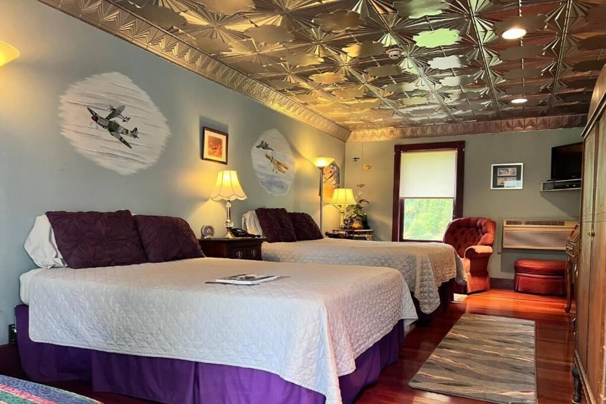A hotel room with two beds, a wooden floor, a ceiling with a geometric pattern, and a window with blinds.