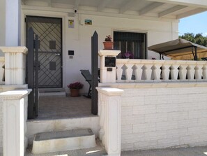 A white building with a gated entrance, steps leading to a porch, a potted plant, and a covered area with a table and chairs. - Mery Giò vacation home in San Pietro in Bevagna suitable for families and groups (San Pietro In Bevagna)
