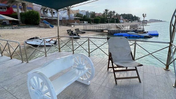 A covered outdoor seating area with a bench and lounge chair, overlooking a beach and water. - Lemon Beach Chalet in Durrat Al Arous (درة العروس)