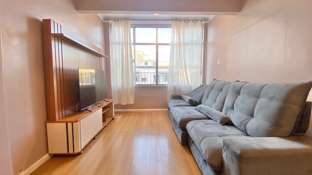 Een woonkamer met een televisie op een houten dressoir, een grijze bank en een raam met gordijnen.
