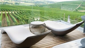 A terrace with a modern lounge chair, a small round table with two glasses, and a view of a vineyard. - Vineho (Barolo)