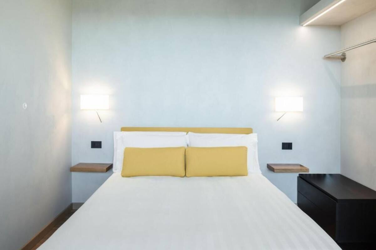 A bed with white and yellow bedding, two bedside tables, and wall-mounted lights.