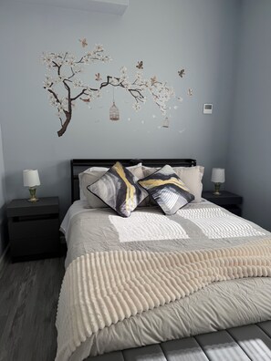 Un dormitorio con cama, dos mesitas de noche, una lámpara fijada a la pared y un adorno de pared con un árbol, flores y mariposas.