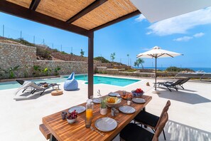 Ein Holztisch, gedeckt für das Frühstück, mit Blick auf einen Swimmingpool und das Meer.