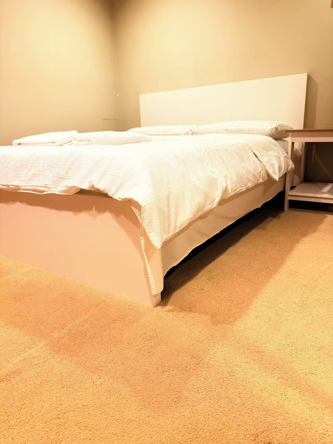 Ein ordentlich bezogenes Bett mit weißem Kopfteil in einem Zimmer mit Teppichboden und einem kleinen Nachttisch.